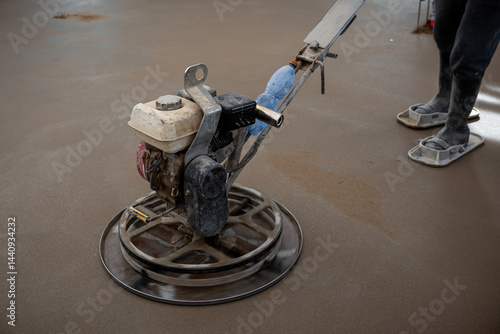 A construction worker operates a power trowel machine indoors, smoothing a freshly laid concrete floor. Protective footwear and active trowel blades are visible.