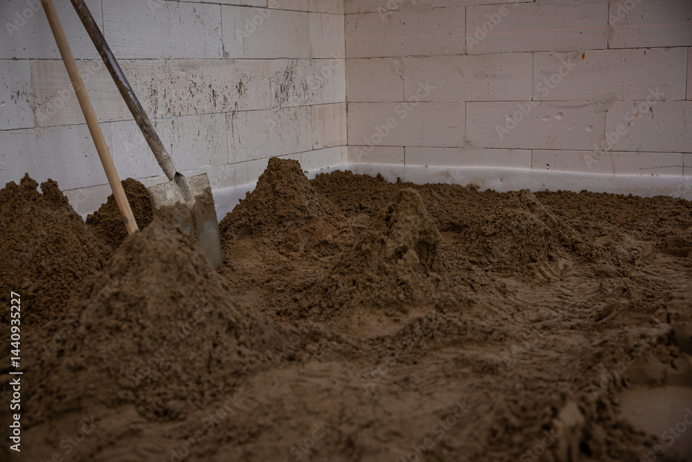 Interior of a construction site with sand piles on the floor, a shovel against white block walls, and visible signs of ongoing work.
