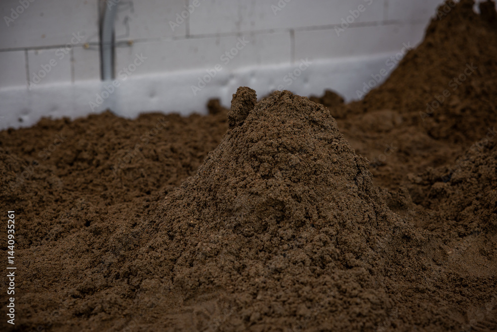 Indoor construction site with a pile of brown soil, a white wall with seams, and exposed piping, prepared for underfloor heating or concrete work.
