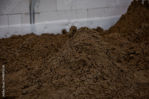 Indoor construction site with a pile of brown soil, a white wall with seams, and exposed piping, prepared for underfloor heating or concrete work.
