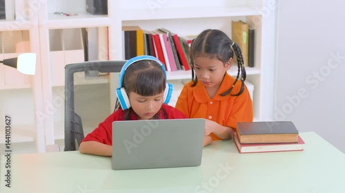 Two young asian girls collaborating on a laptop for online education, wearing headphones and glasses, with books stacked nearby, embodying remote learning and teamwork