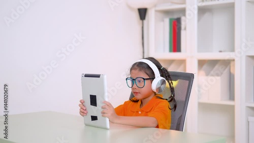 Asian student girl wearing headphones and eyeglasses using tablet computer studying online e-learning system at home, homeschooling, technology and education concept