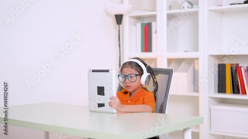 Asian student girl wearing headphones and eyeglasses using tablet computer studying online e-learning system at home, homeschooling, technology and education concept