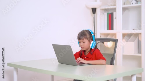 Asian girl studying online using laptop and wearing headphones sitting at desk in her room with bookshelf in background, homeschooling concept
