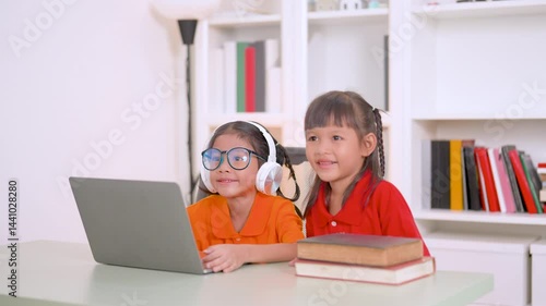 Two young asian girls collaborating on a laptop for online education, wearing headphones and glasses, with books stacked nearby, embodying remote learning and teamwork