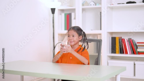 Healthy asian child enjoying a refreshing glass of pure water while sitting on a chair at home, emphasizing the importance of hydration and promoting a vibrant lifestyle