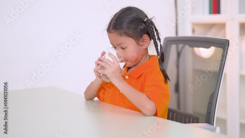 Healthy asian child enjoying a refreshing glass of pure water while sitting on a chair at home, emphasizing the importance of hydration and promoting a vibrant lifestyle