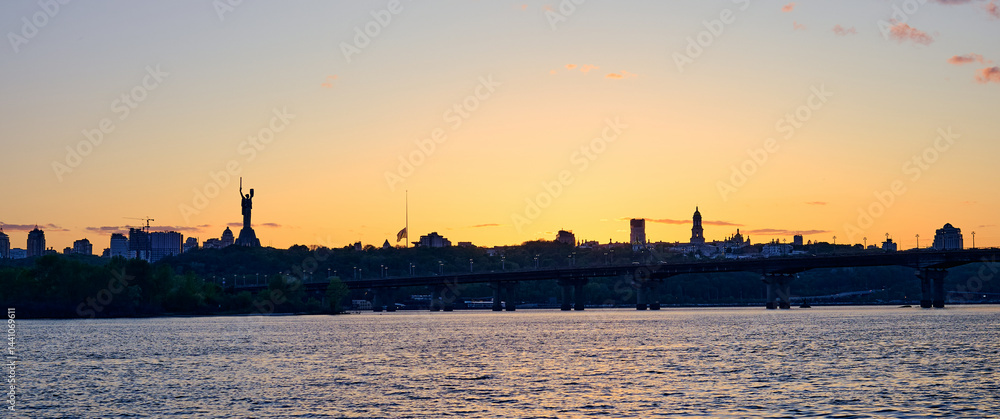 Fototapeta premium Beautiful sunset over Kyiv city skyline with Dnipro River, Paton Bridge, and Motherland Monument silhouette under colorful sky