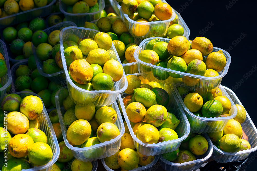 Vibrant assortment of lemons and limes packed closely in plastic containers, highlighting their contrasting yellow and green. Stacked containers overflowing with ripe citrus fruits at street market