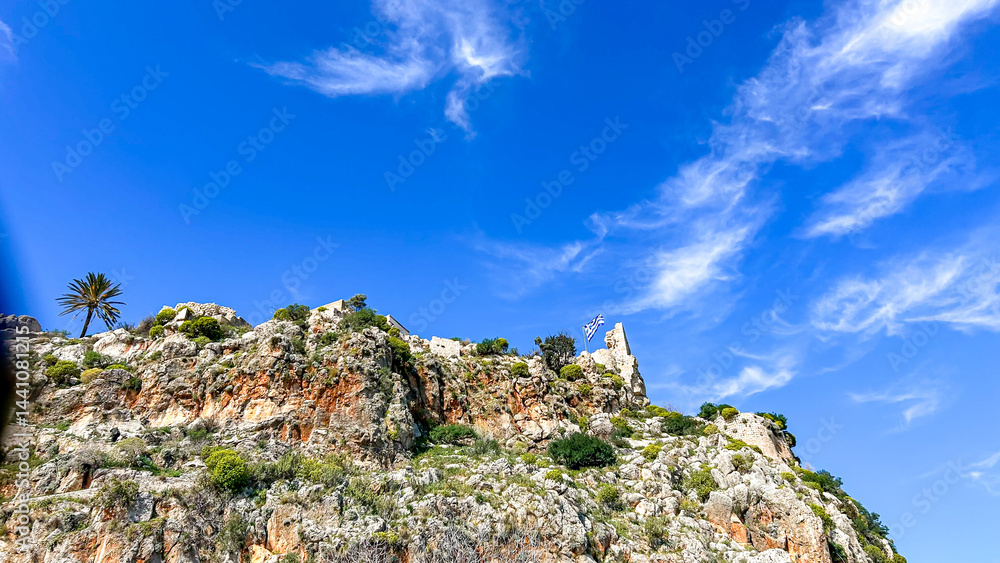 Naklejka premium Ancient Stone Ruins With Greek Flag On Rocky Hilltop In Greece Under Bright Blue Sky, Concept Of Heritage, Summer Travel, And Historical Tourism