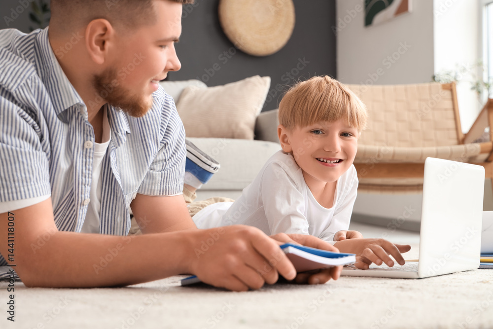 Young man and his little son doing homework while lying on floor with laptop in living room Young man and his little son doing homework while lying on floor with laptop in living room