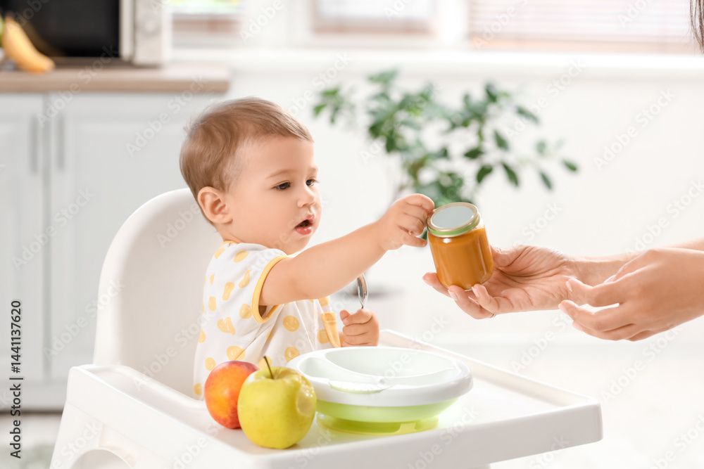 Mother with jars of tasty food and fruits feeding cute little baby on chair in kitchen at home