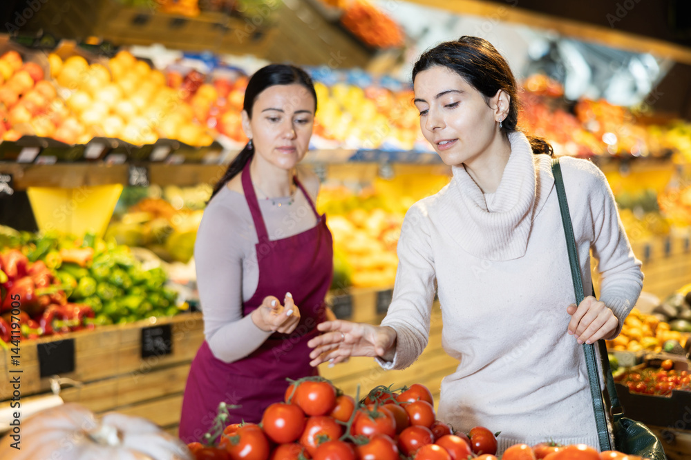 Obraz premium Young woman buyer choosing fresh tomatoes with help of adult woman seller in vegetable shop