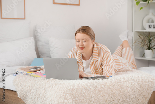 Beautiful young happy woman in pajamas working with laptop on bed at home. Re...