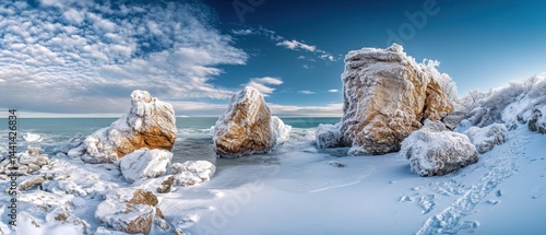 Panoramic vista of a serene winter landscape featuring snow covered rocks along a frozen coastline under a bright blue sky with scattered clouds