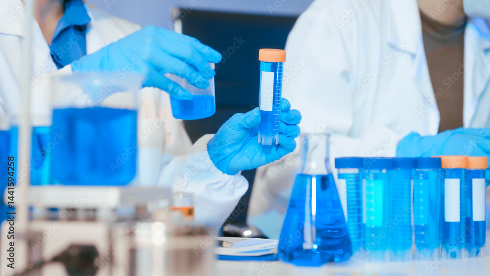 Two Asian female scientists wearing laboratory gloves sit at a table, carefully examining a test tube filled with blue liquid, using a microscope for detailed analysis in a research laboratory Two Asian female scientists wearing laboratory gloves sit at a table, carefully examining a test tube filled with blue liquid, using a microscope for detailed analysis in a research laboratory