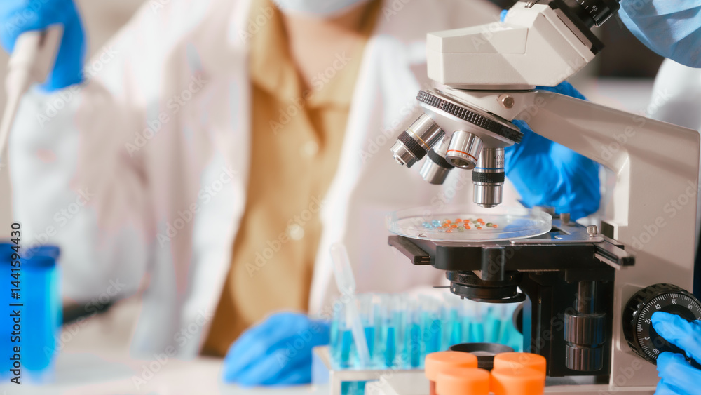 Two Asian female scientists wearing laboratory gloves sit at a table, carefully examining a test tube filled with blue liquid, using a microscope for detailed analysis in a research laboratory