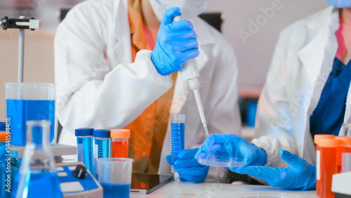 Two Asian female scientists wearing laboratory gloves sit at a table, carefully examining a test tube filled with blue liquid, using a microscope for detailed analysis in a research laboratory