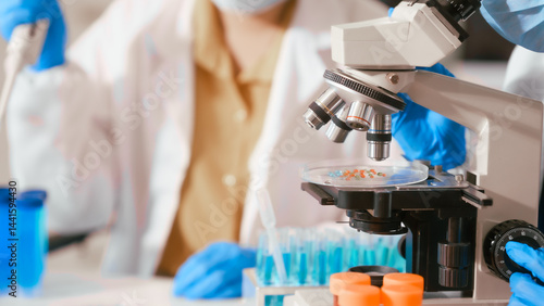 Obraz na płótnie Two Asian female scientists wearing laboratory gloves sit at a table, carefully examining a test tube filled with blue liquid, using a microscope for detailed analysis in a research laboratory w salonie