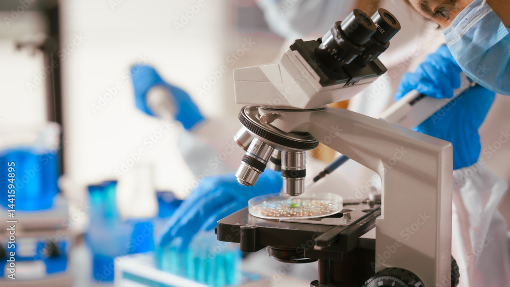 Two Asian female scientists wearing laboratory gloves sit at a table, carefully examining a test tube filled with blue liquid, using a microscope for detailed analysis in a research laboratory