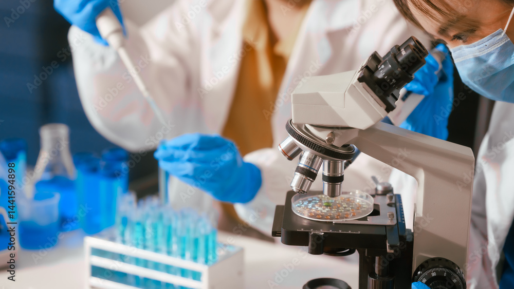 Two Asian female scientists wearing laboratory gloves sit at a table, carefully examining a test tube filled with blue liquid, using a microscope for detailed analysis in a research laboratory