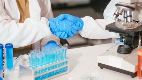 Two Asian female scientists wearing laboratory gloves sit at a table, carefully examining a test tube filled with blue liquid, using a microscope for detailed analysis in a research laboratory