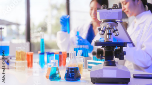 Obraz na płótnie Two Asian female scientists wearing laboratory gloves sit at a table, carefully examining a test tube filled with blue liquid, using a microscope for detailed analysis in a research laboratory w salonie