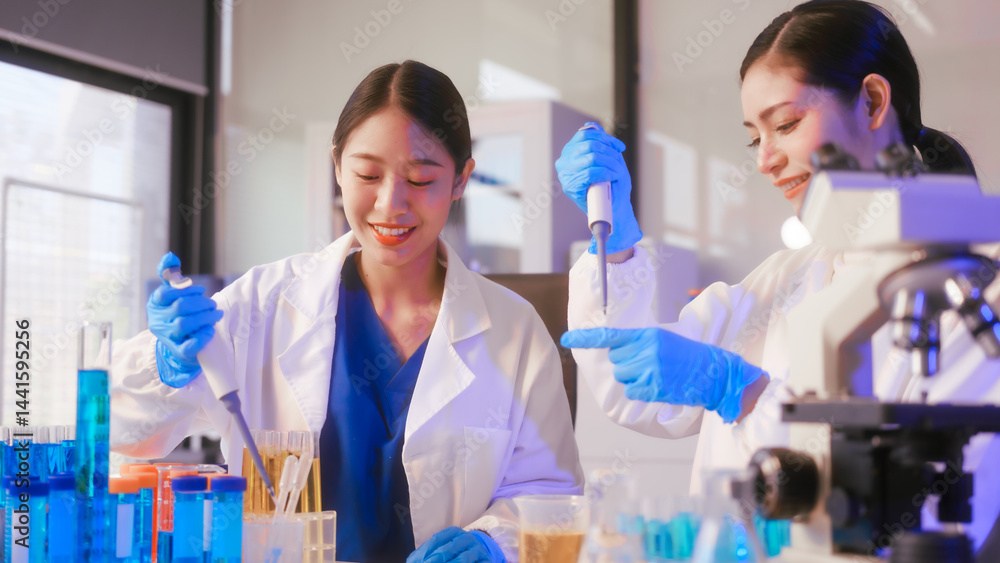 Two Asian female scientists wearing laboratory gloves sit at a table, carefully examining a test tube filled with blue liquid, using a microscope for detailed analysis in a research laboratory