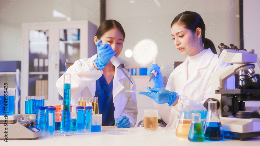 Two Asian female scientists wearing laboratory gloves sit at a table, carefully examining a test tube filled with blue liquid, using a microscope for detailed analysis in a research laboratory