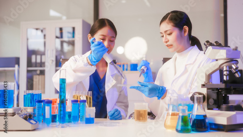 Obraz na płótnie Two Asian female scientists wearing laboratory gloves sit at a table, carefully examining a test tube filled with blue liquid, using a microscope for detailed analysis in a research laboratory w salonie