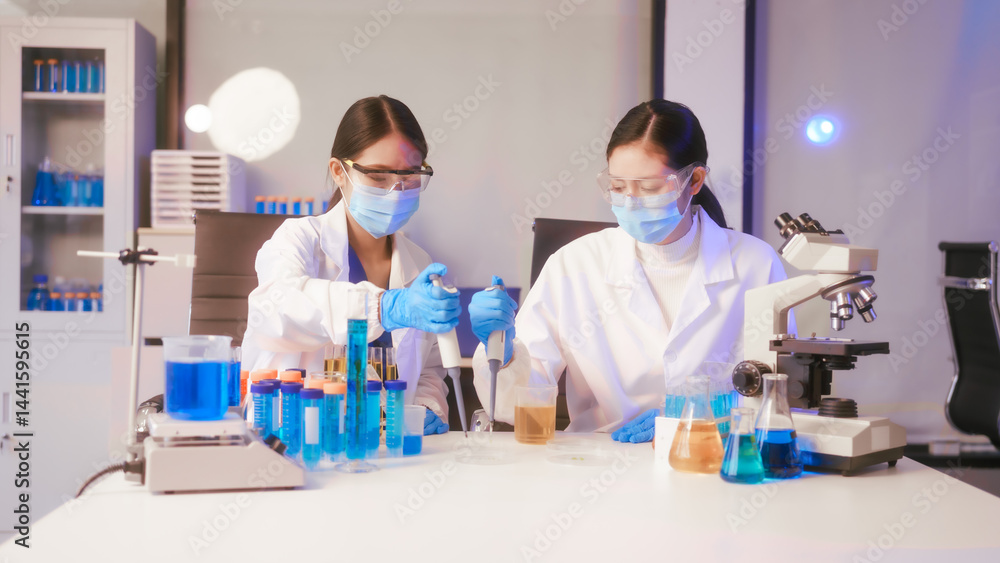 Two Asian female scientists wearing laboratory gloves sit at a table, carefully examining a test tube filled with blue liquid, using a microscope for detailed analysis in a research laboratory