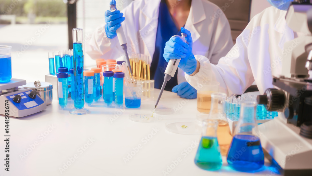 Two Asian female scientists wearing laboratory gloves sit at a table, carefully examining a test tube filled with blue liquid, using a microscope for detailed analysis in a research laboratory
