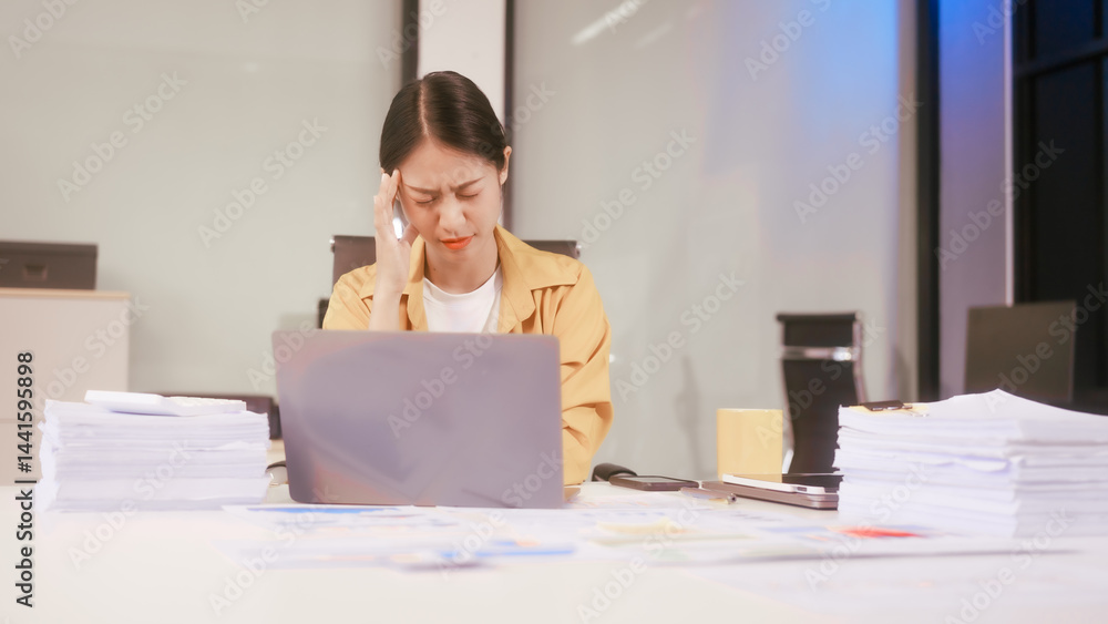 An Asian woman works at her office desk, feeling stressed and experiencing pain from sitting for a long time, struggling with discomfort and fatigue while trying to stay focused on her tasks.