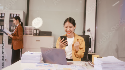 A young Asian businesswoman works at her office table, smiling happily while looking at her phone, feeling joyful and proud of her success in achieving professional goals in the workplace