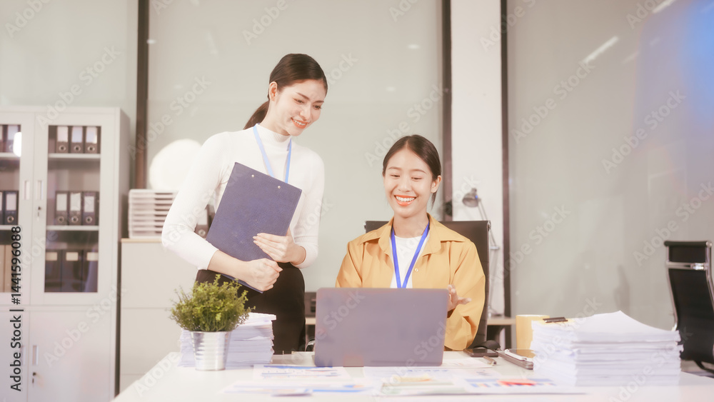 Two young Asian business woman sit at a table in the office, discussing marketing strategies, using a computer reviewing documents with charts and graphs, collaborating on new business plans at work