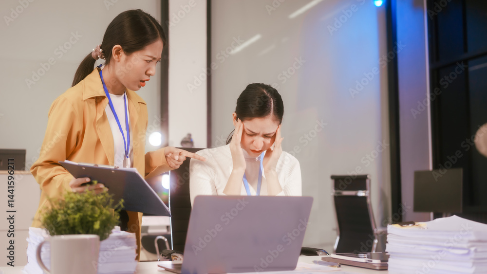 Two Asian women sit working at desk,overwhelmed with stress struggling with headaches,feeling mentally and physically exhausted while trying to concentrate on their demanding tasks in a busy office