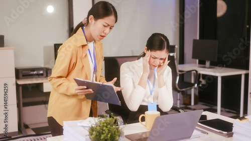 Two Asian women sit working at  desk,overwhelmed with stress struggling with headaches,feeling mentally and physically exhausted while trying to concentrate on their demanding tasks in a busy office