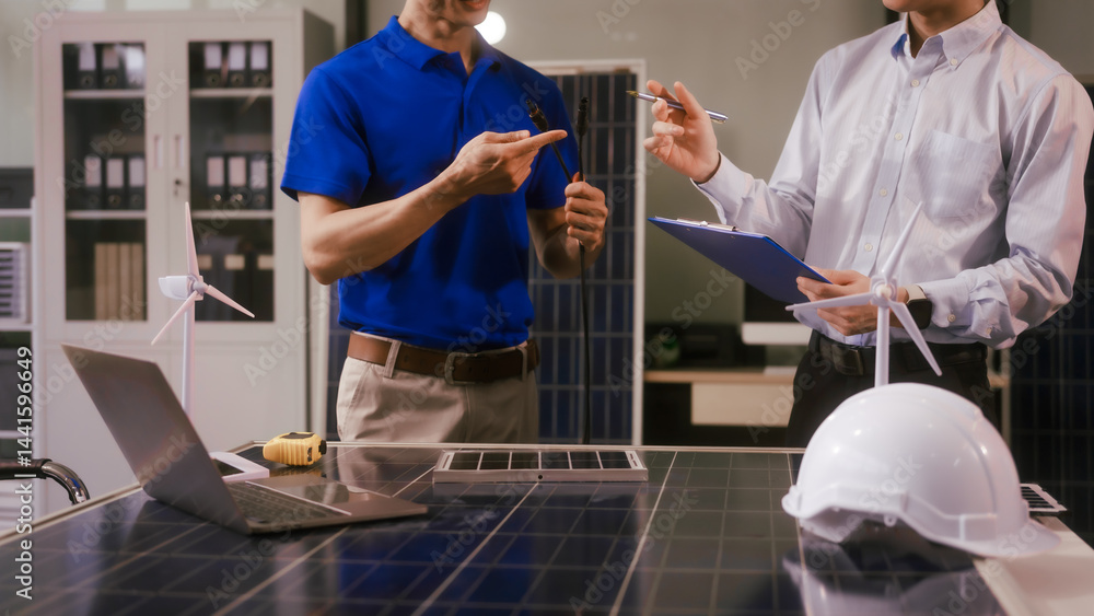 Two Asian businessmen wearing engineer hats table, discussing solar panels and wind turbines. They plan environmental projects, focusing on solar energy solutions and renewable energy installations