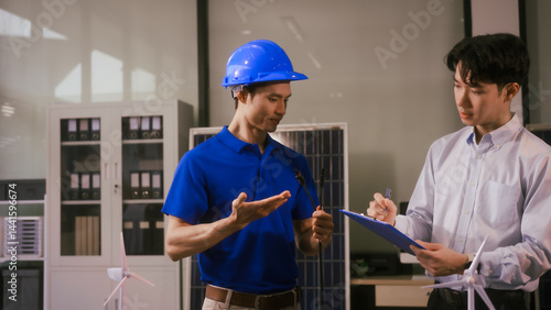Obraz na płótnie Two Asian businessmen wearing engineer hats table, discussing solar panels and wind turbines. They plan environmental projects, focusing on solar energy solutions and renewable energy installations w salonie