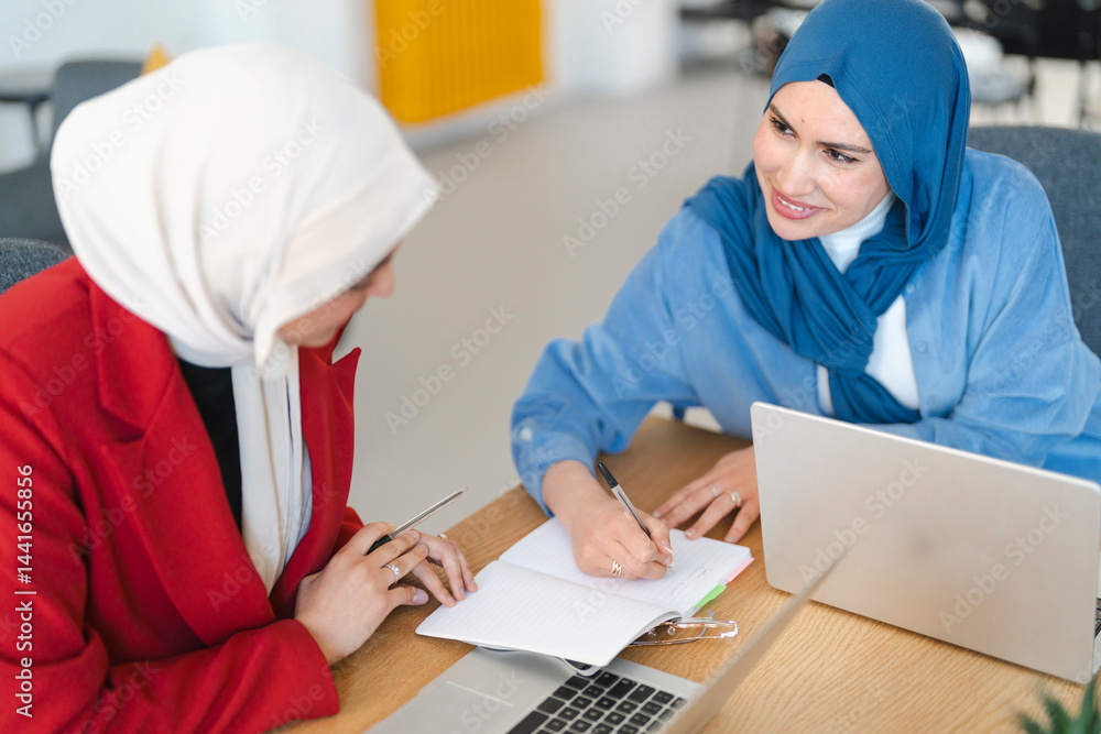 Two women in hijabs collaborate on a project. They're brainstorming ideas and taking notes, working together to achieve a common goal. Two women in hijabs collaborate on a project. They're brainstorming ideas and taking notes, working together to achieve a common goal.