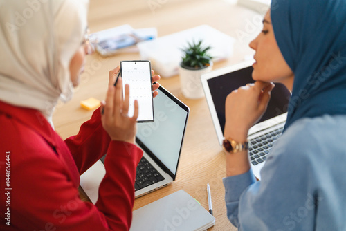Two women in hijabs are collaborating on a project. They're using laptops and a calendar app to schedule and plan their work, ensuring they stay organized and on track.