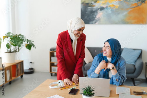 Two smiling women in hijabs collaborate on a project at a desk. They're likely working together on a business or creative endeavor, enjoying the process and each other's company.