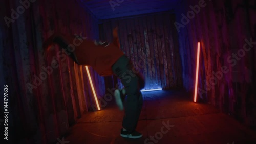 Full shot of young talented Caucasian b-boy performing breakdance indoors under blue and orange ambient light showing excellent footwork and creativity