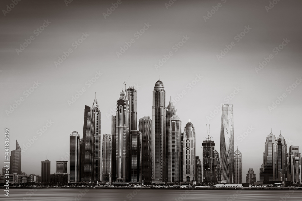 Fototapeta premium Monochrome city skyline with tall skyscrapers reflecting on calm water under a cloudy sky, Dubai Marina bay UAE