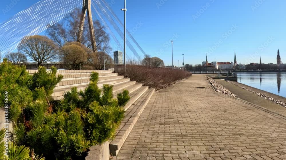 Scenic view of the Vansu Bridge from Kipsala Island in Riga, Latvia, with the cityscape in the background, featuring a paved promenade, lush greenery, and concrete steps, on a bright sunny day Scenic view of the Vansu Bridge from Kipsala Island in Riga, Latvia, with the cityscape in the background, featuring a paved promenade, lush greenery, and concrete steps, on a bright sunny day