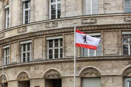 Photography The Berlin state flag with the iconic bear emblem waves in front of a historic stone building facade with ornate details and classic European architecture