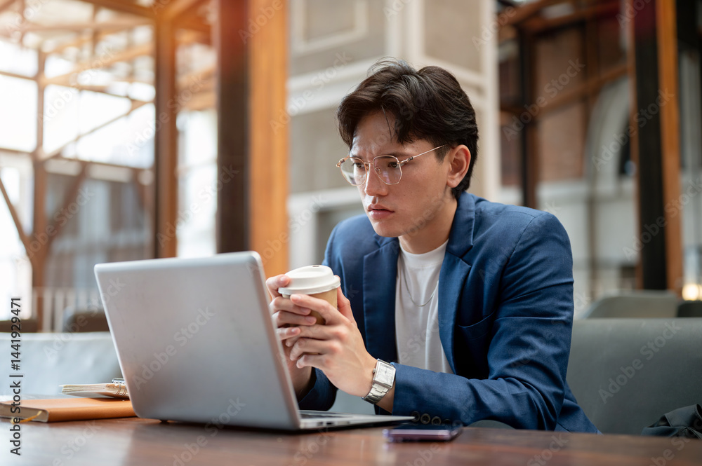 Blue suit asian man is looking troubled while holding coffee cup and looking at his laptop on table. Blue suit asian man is looking troubled while holding coffee cup and looking at his laptop on table.
