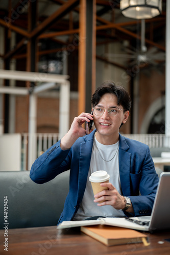 Blue suit man talking on phone while holding coffee cup, sitting with laptop in cafe or coffee shop.