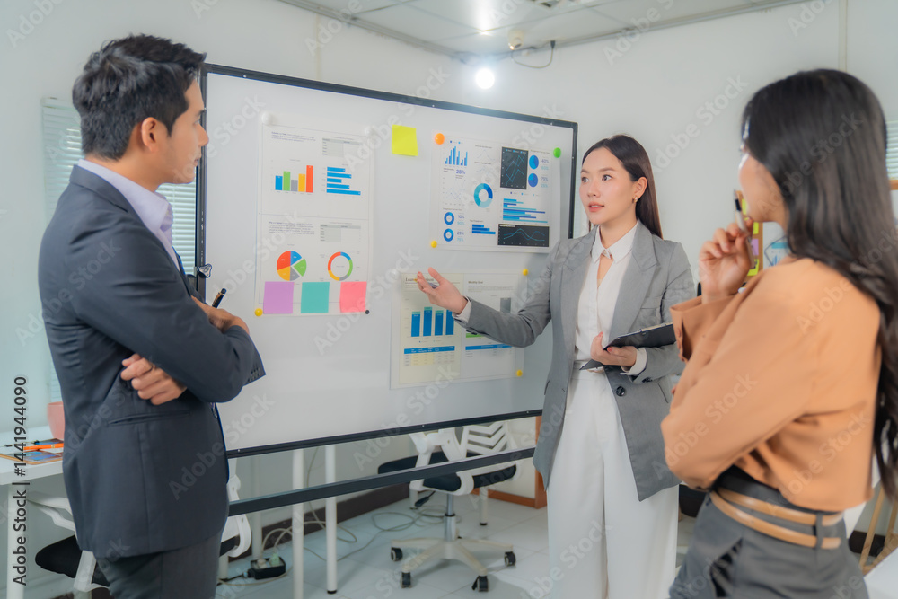 Fototapeta premium Three Asian business professionals engaging in a strategic discussion during a meeting in a modern office, utilizing a whiteboard filled with insightful charts and graphs