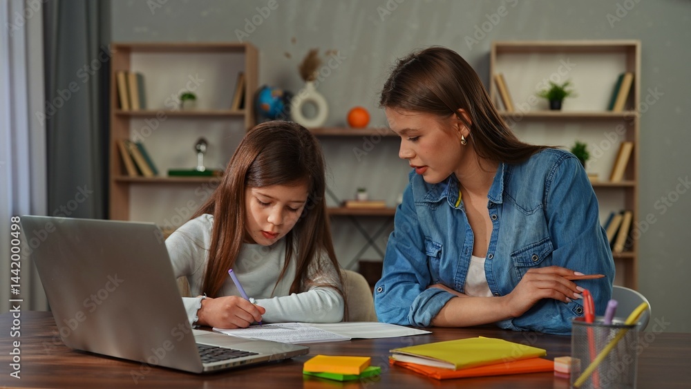 Obraz premium Young child girl studying at home, small girl sits at desk and attends school class online on laptop, doing homework, mother helping and controlling process.